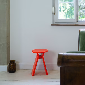 Ein leuchtend roter Hocker mit dem Namen "Koshi" steht in einer minimalistisch eingerichteten Ecke eines Raumes mit weißem Holzboden. Daneben eine braune Vase, im Hintergrund ein Fenster mit Blick ins Grüne und ein Teil eines rustikalen Sofas.
