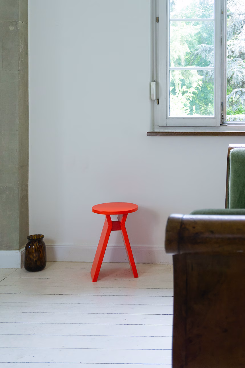 Ein leuchtend roter Hocker mit dem Namen "Koshi" steht in einer minimalistisch eingerichteten Ecke eines Raumes mit weißem Holzboden. Daneben eine braune Vase, im Hintergrund ein Fenster mit Blick ins Grüne und ein Teil eines rustikalen Sofas.
