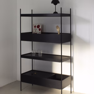 Das schwarze Metallregal "Felo" von Forma steht in einem schlichten, hellen Raum. Es hat vier Ablagen, auf denen sich eine kleine Lampe, ein Buch mit roten Punkten, eine Holzhand, zwei Wassergläser und dekorative Kreisel befinden.